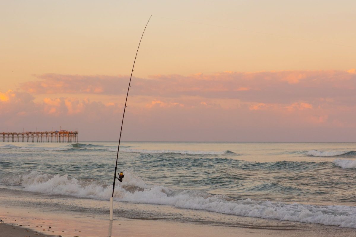 A peaceful sunset scene featuring a surf fishing rod planted in the sand as waves gently crash along the shore. With a coastal pier visible in the distance and soft pink clouds overhead, this image captures the relaxing outdoor lifestyle enjoyed by guests of Coastline Realty Vacations.